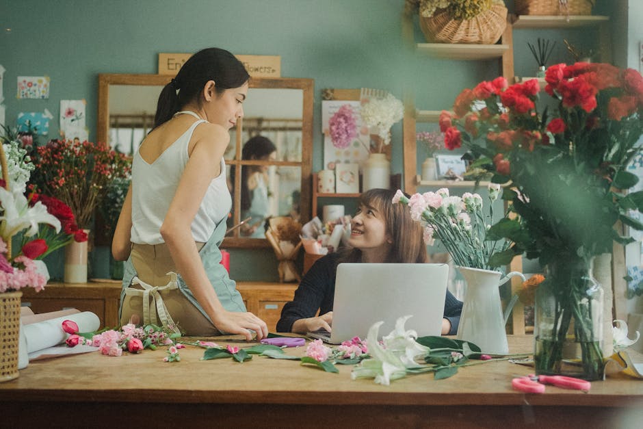 A young female florist with dark hair, wearing a white sleeveless top and beige apron, stands at a wooden workspace inside a flower shop. She is leaning slightly over the table, engaging in conversati