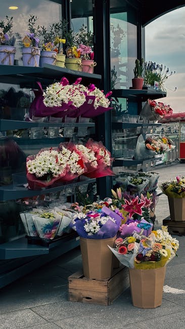 A display of various floral arrangements and bouquets outside a florist shop, featuring vibrant pink, white, purple, and yellow flowers arranged in paper and fabric wrapping, placed on shelves, wooden