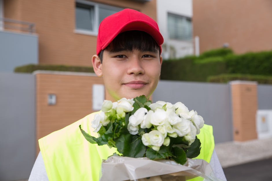 A young male flower delivery courier wearing a red cap and high-visibility jacket is holding a bouquet of fresh white roses wrapped in white paper with green leaves visible. He stands outdoors in fron