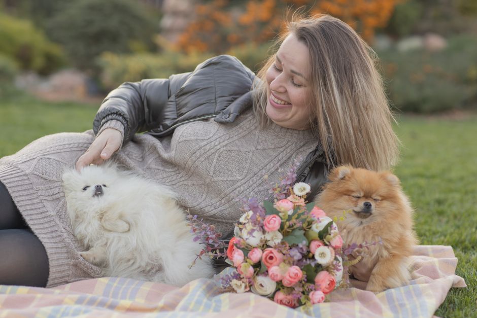 A woman with light brown, shoulder-length hair lying on a colorful checkered blanket outdoors in a park or garden setting. She is smiling gently, wearing a grey knitted sweater and a grey puffer jacke