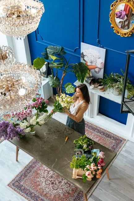 A woman in a beige top and dark skirt arranging a bouquet of fresh flowers on a grey table inside a vibrant blue room. The floral arrangement includes white lilies, pink roses, purple lilacs, and gree